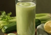 Painterly realistic glass of zucchini, cucumber, celery, and lemon green juice on a wooden board with fresh vegetables and linen cloth